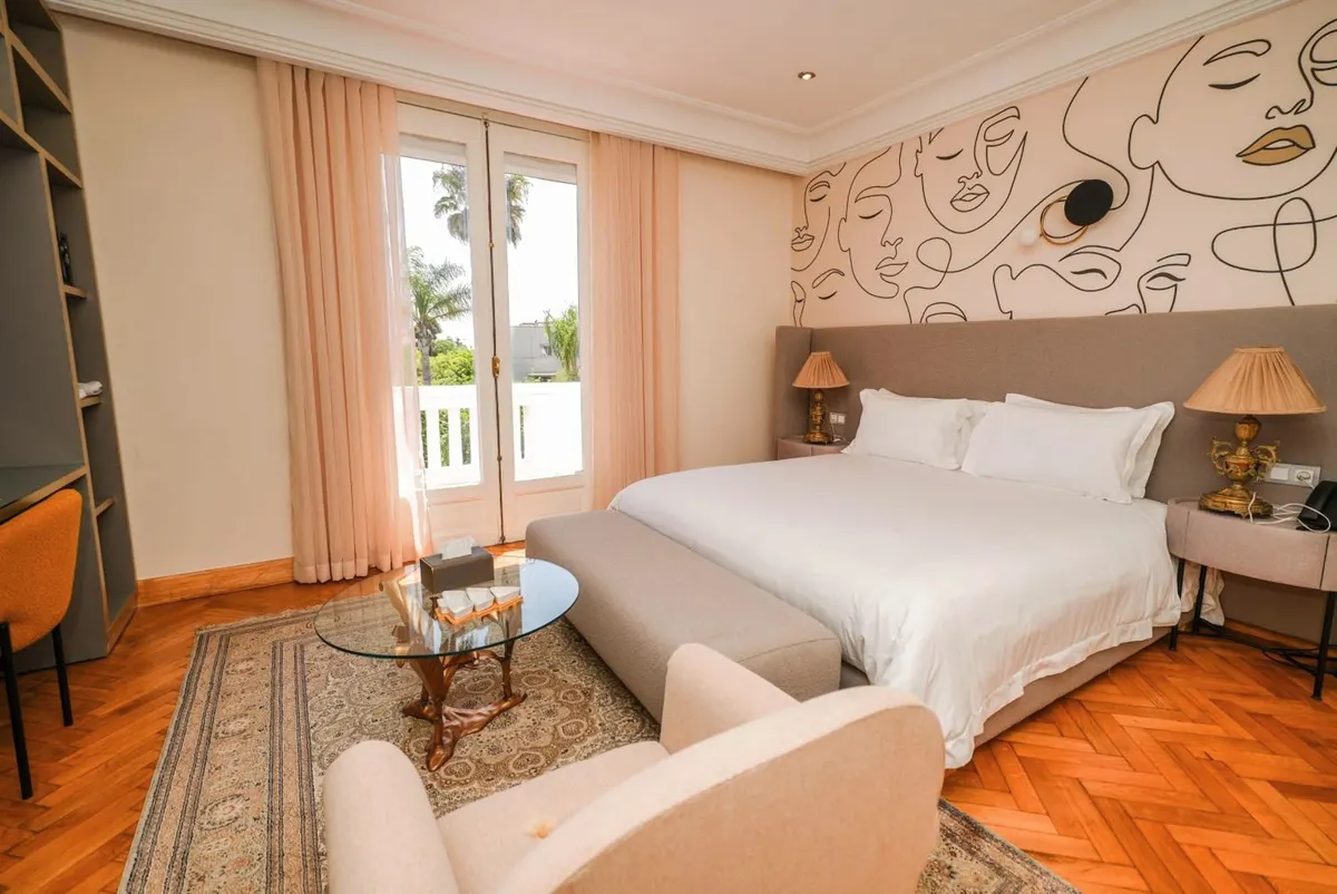 Deluxe Double Room: bed against a line-drawn mural of faces, armchairs and French windows to the balcony.